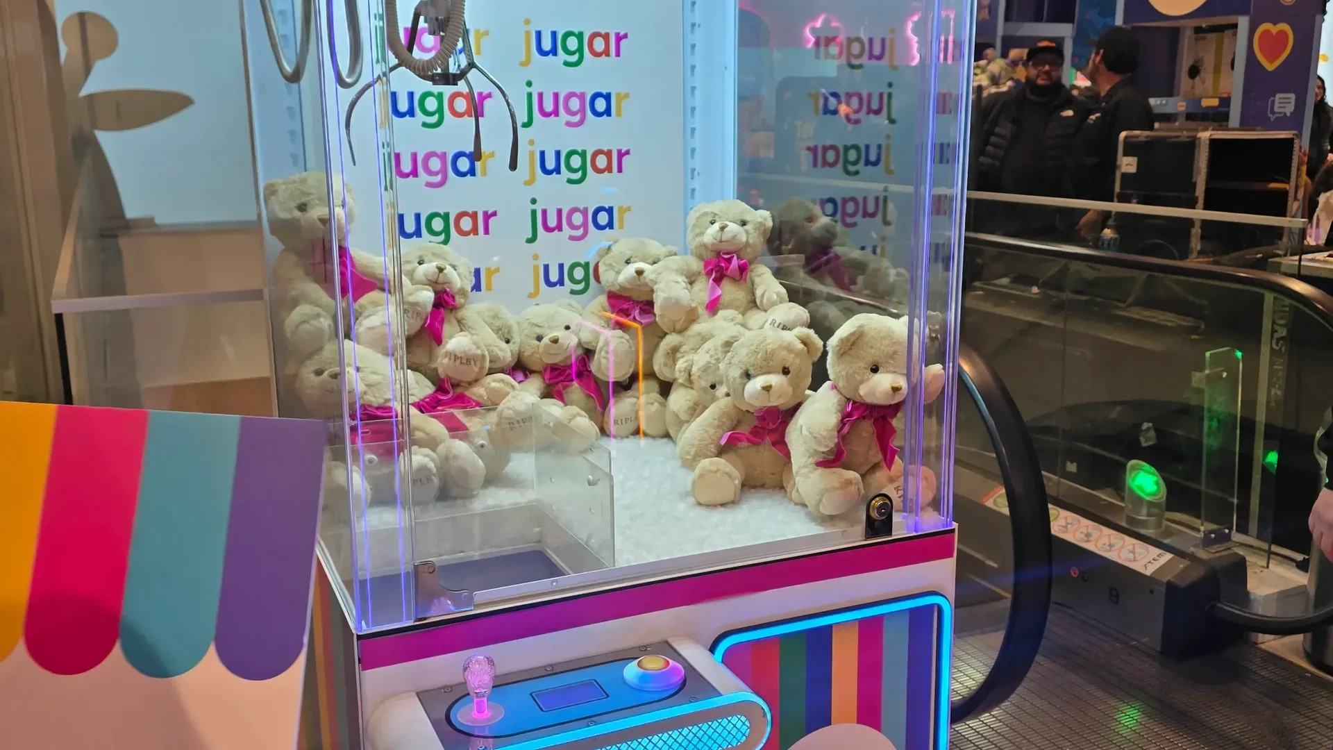 Máquina atrapa peluches rosada de Ripley llena de ositos de peluche con lazos, iluminada con neón en un mall